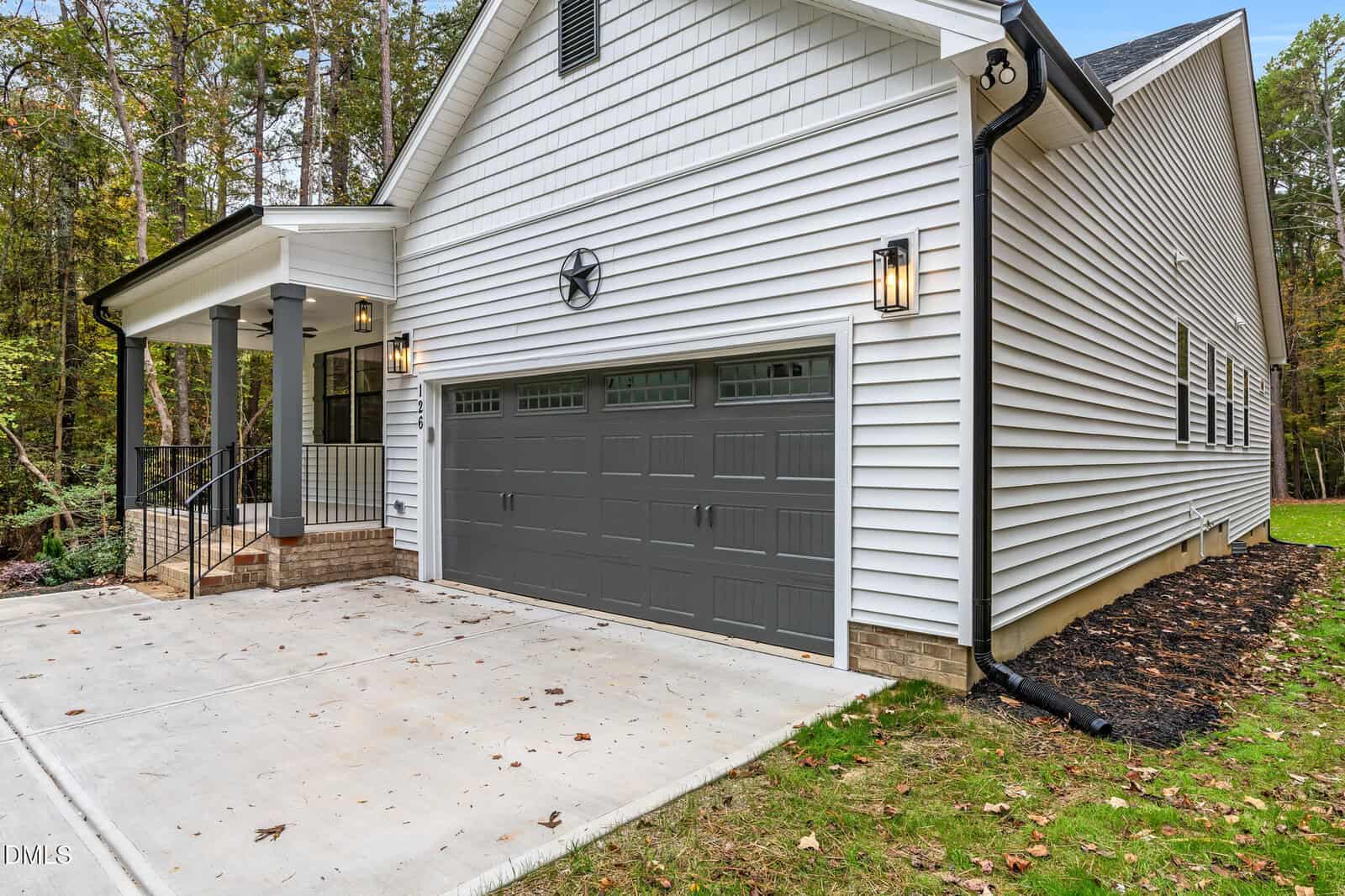 A modern house with white siding, a gray double garage door, black outdoor lights, a star decoration above the garage, a small covered porch with steps and railings, and a concrete driveway. Trees are visible in the background.