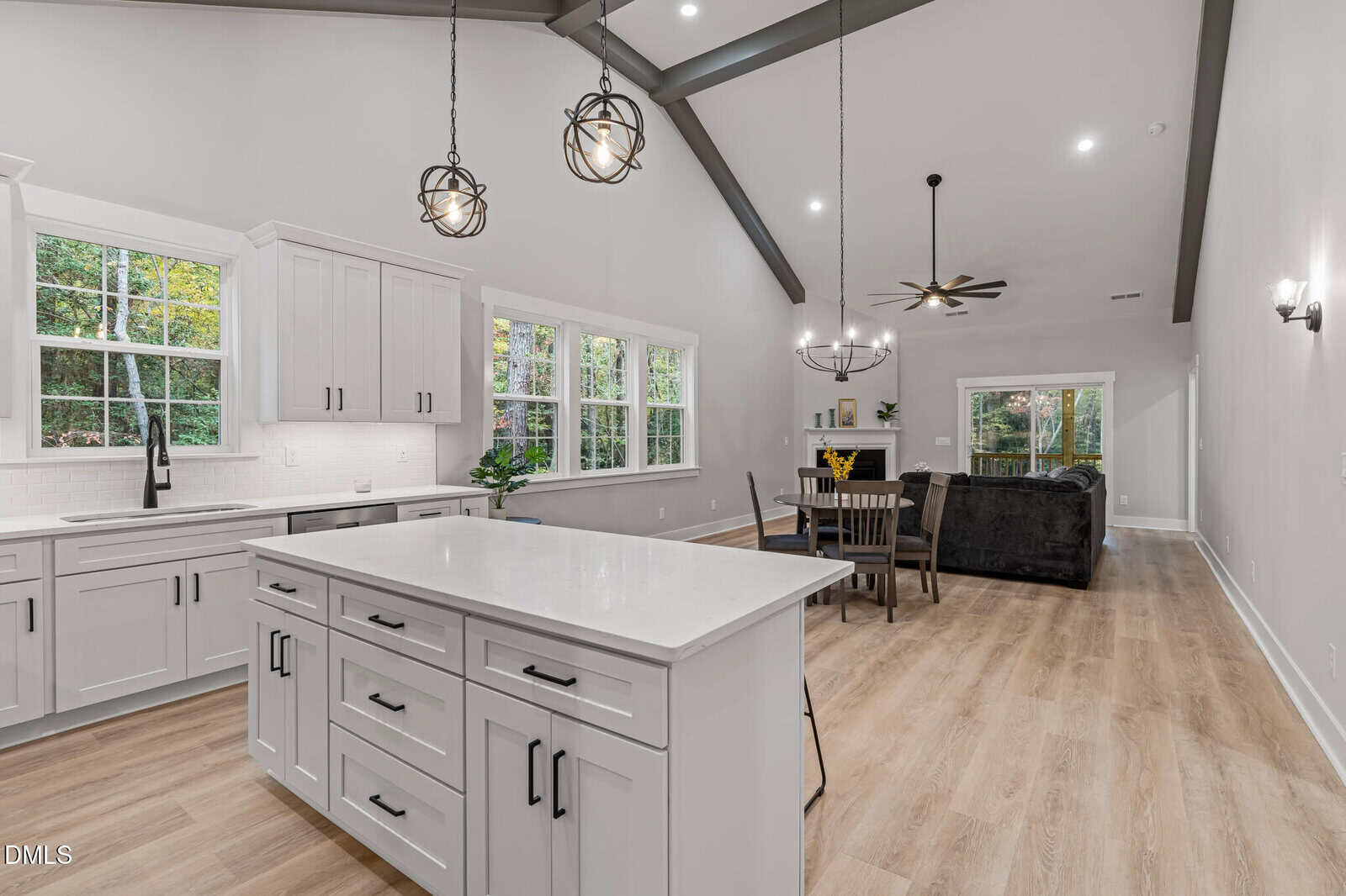 Modern open-concept kitchen and living area with white cabinets, a large island, wood floors, and two pendant lights. The dining table seats six, and there is a black sofa set near large windows overlooking a green outdoor view.