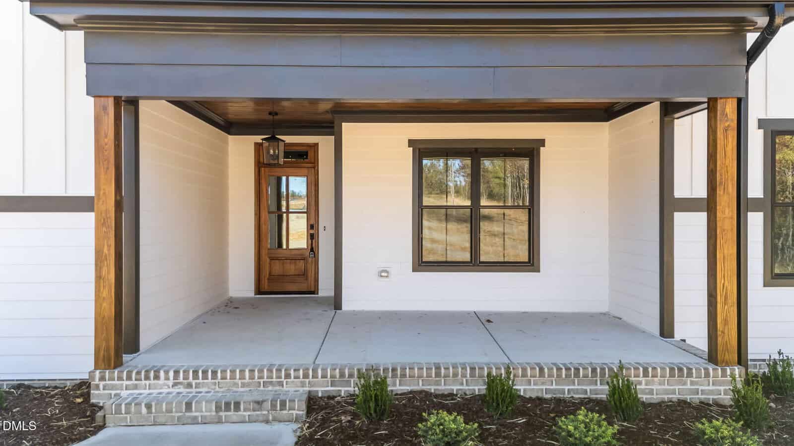 A covered front porch with wood posts, a wooden front door with glass panels, a single window, white siding, and a small landscaped area with shrubs in front of a brick-lined concrete porch.