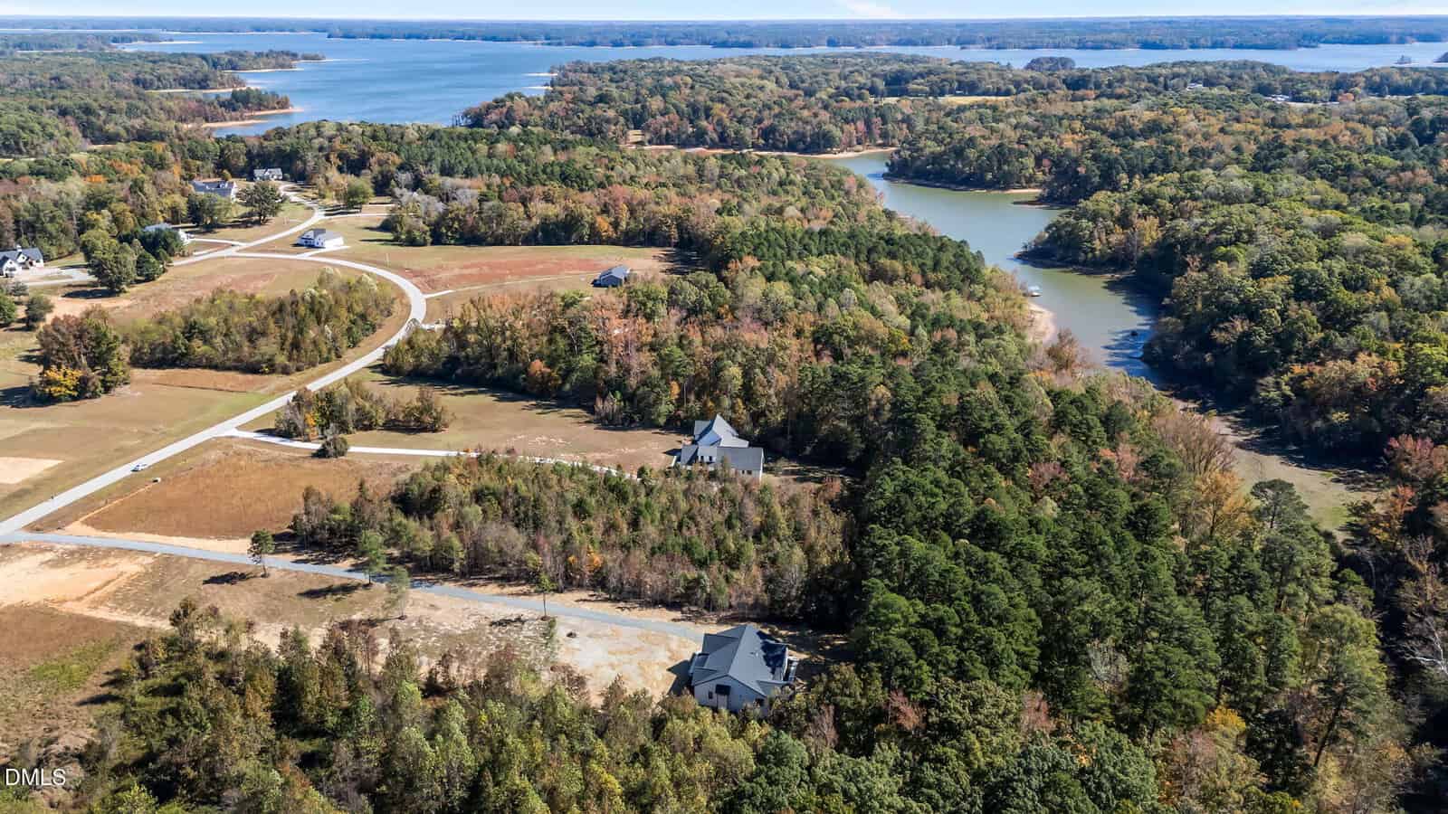Aerial view of houses surrounded by trees and greenery near a winding river and a large lake, with roads connecting homes and forested areas under a clear sky.