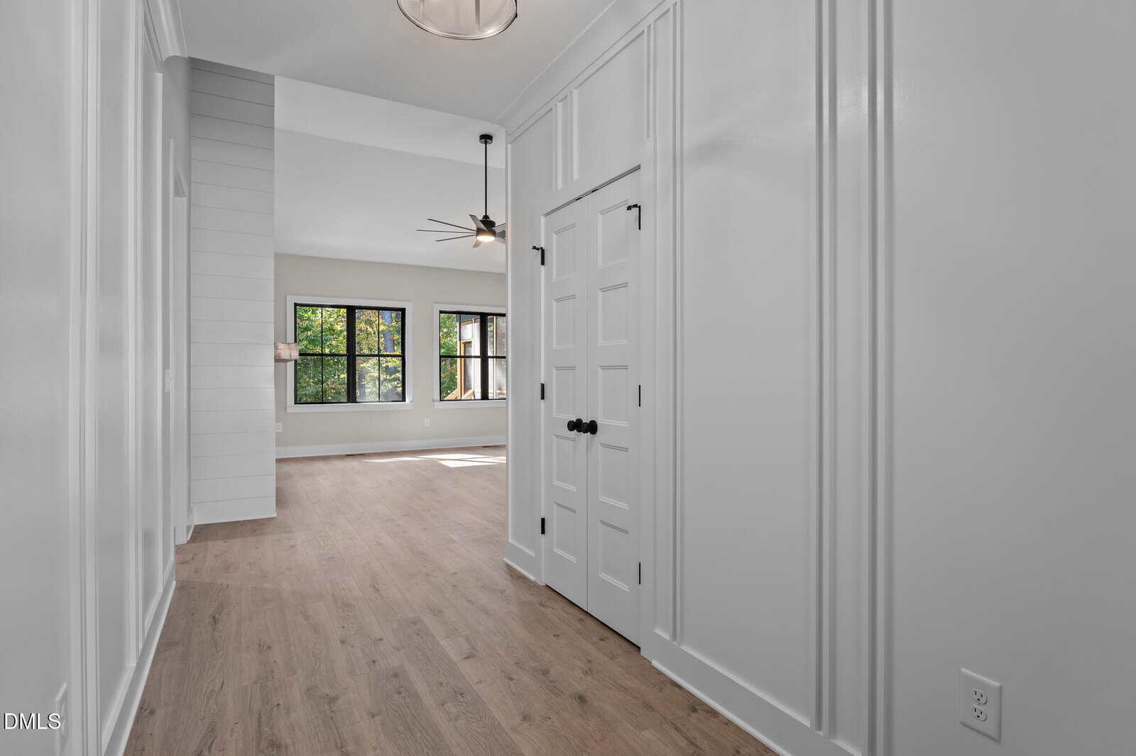 A hallway with light wood flooring and white paneled walls leads to a bright living area with large black-framed windows, a ceiling fan, and modern light fixtures. Double doors are on the right side of the hallway.