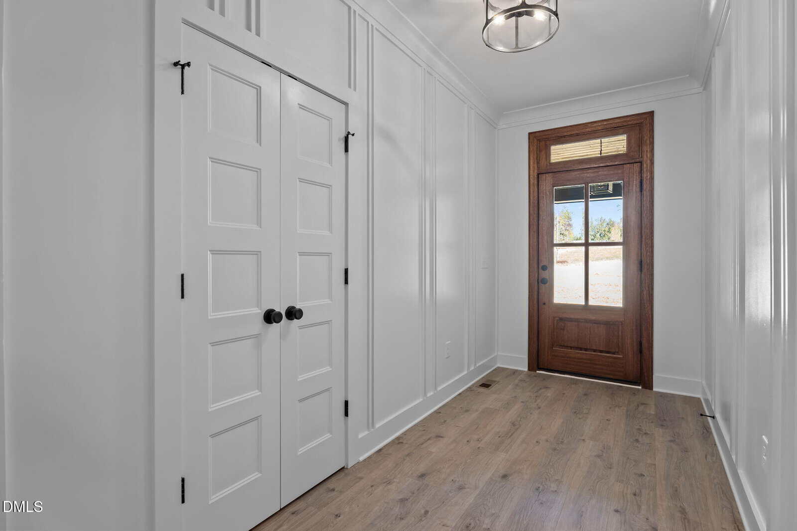 A hallway with light wood floors, white paneled walls, a double white closet door with black knobs on the left, and a wooden front door with glass panels letting in natural light.