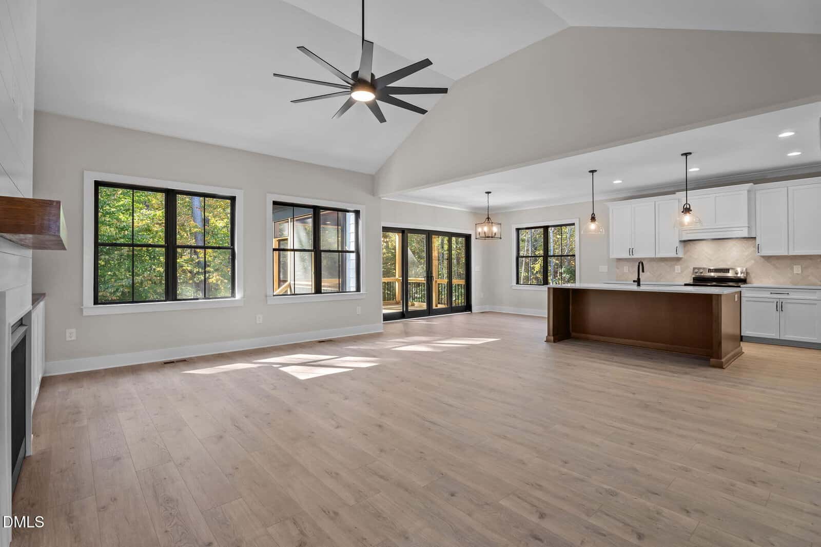 A spacious, empty open-plan living area with light wood flooring, large windows, a fireplace, a central kitchen island, white cabinetry, modern pendant lights, and sliding glass doors to the outside.