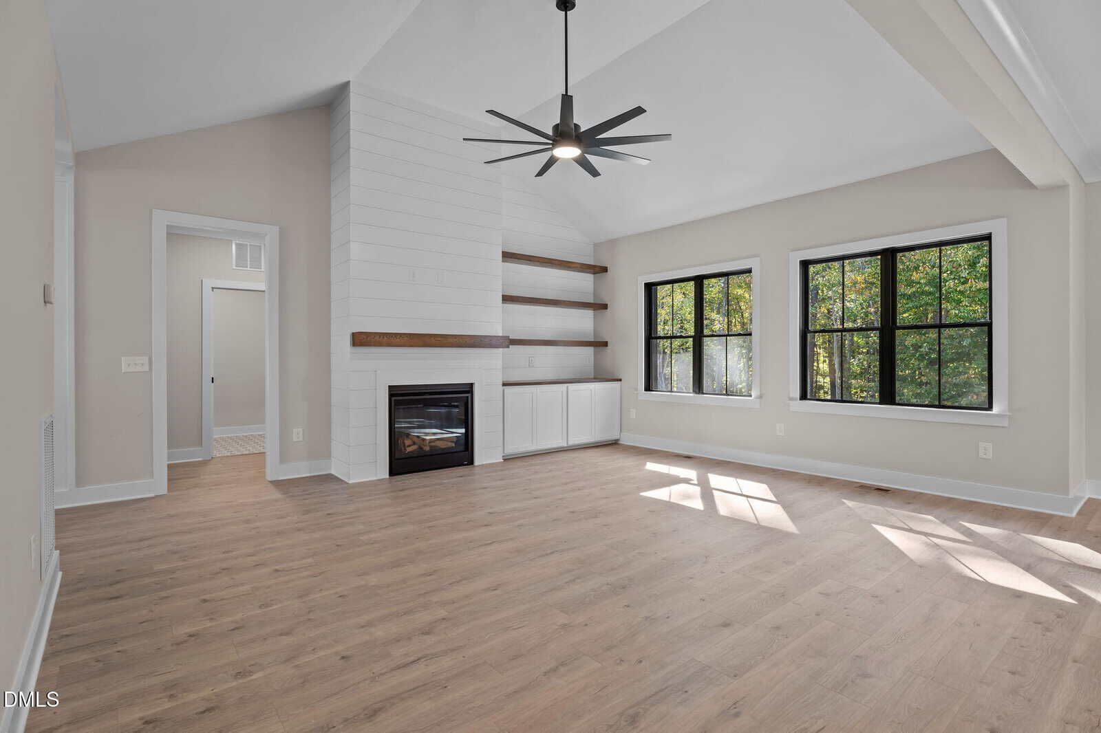 A bright, empty living room with light wood flooring, white walls, black-framed windows, a modern ceiling fan, a white shiplap fireplace, and built-in wooden shelves and cabinets. Sunlight streams in through the windows.