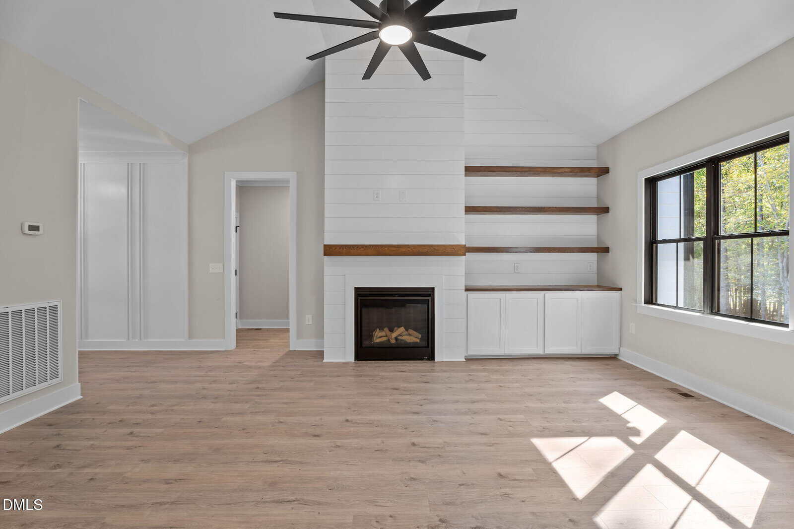 Bright living room with light wood floors, built-in shelves, a white fireplace, large window with black trim, and a modern black ceiling fan. Sunlight streams in, highlighting the clean, minimalist design.