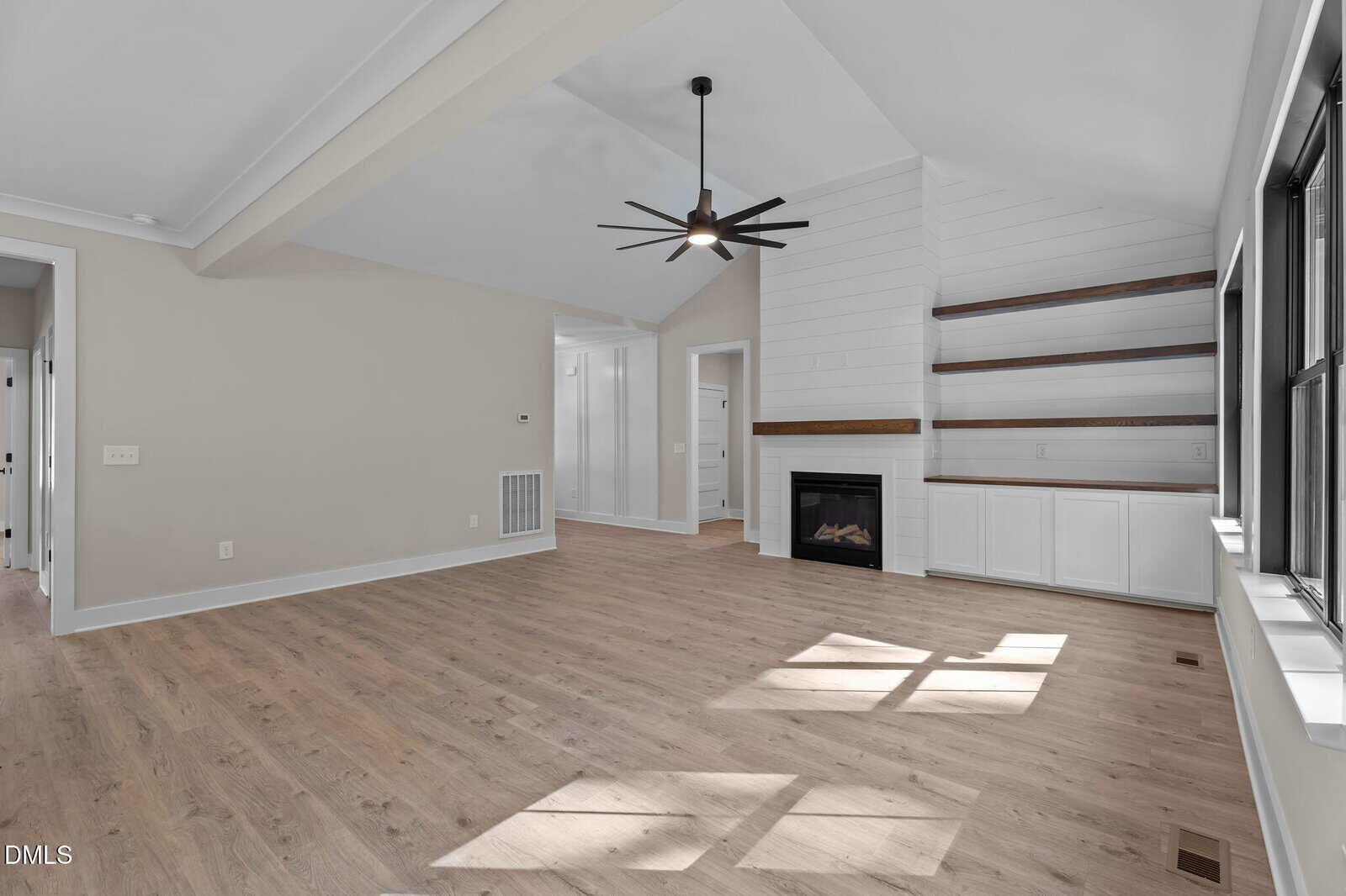 A modern, empty living room with light wood floors, beige walls, a black ceiling fan, a gas fireplace, built-in white shelves and cabinets, and large windows letting in natural light.