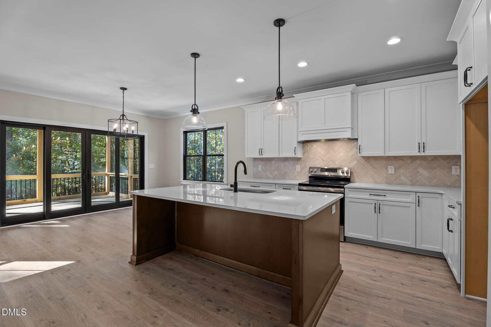 Modern kitchen with white cabinets, stainless steel appliances, a dark island with white countertop, pendant lights, wood floors, and large windows and glass doors opening to a deck with trees outside.