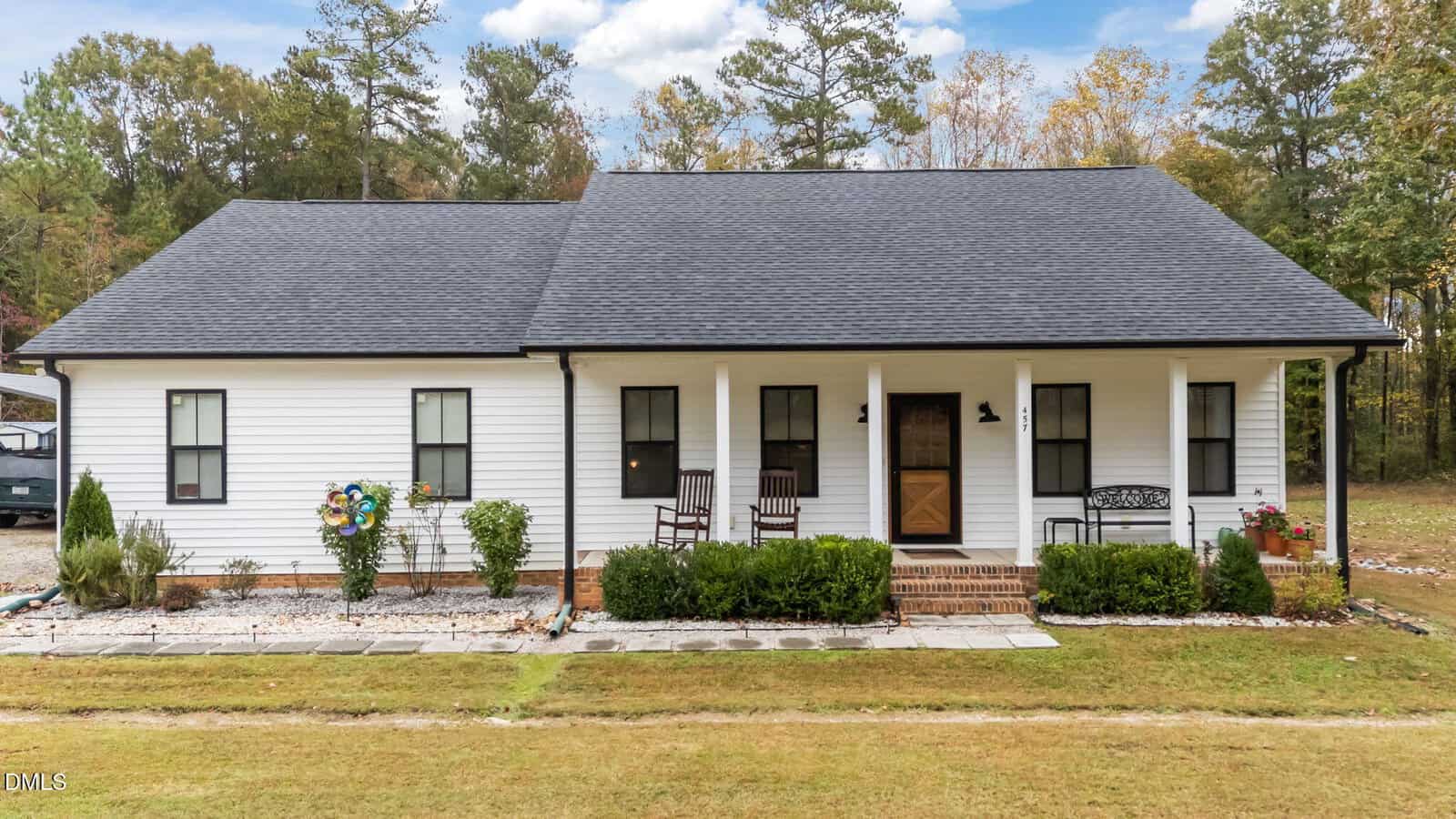 Single-story white house with a black roof and covered front porch, featuring two rocking chairs, a bench, shrubs, a flower decoration, and a stone walkway in a grassy yard with trees in the background.