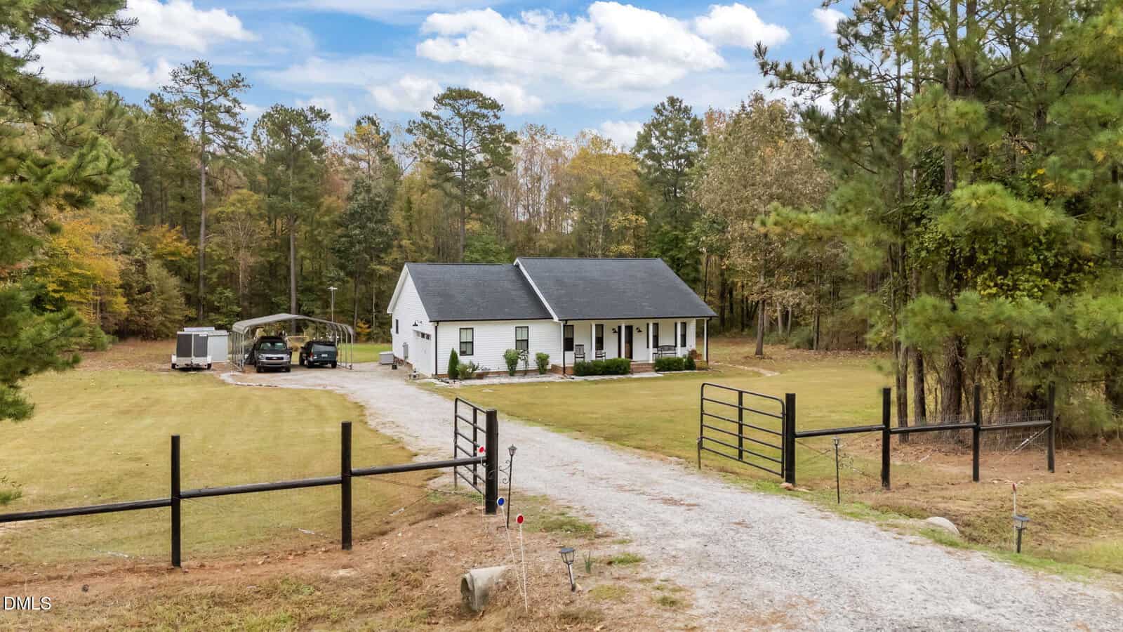 A single-story white house with a black roof sits in a large, grassy yard surrounded by trees. There is a gated gravel driveway, a carport with two vehicles, and a trailer to the left of the house under a blue sky with clouds.