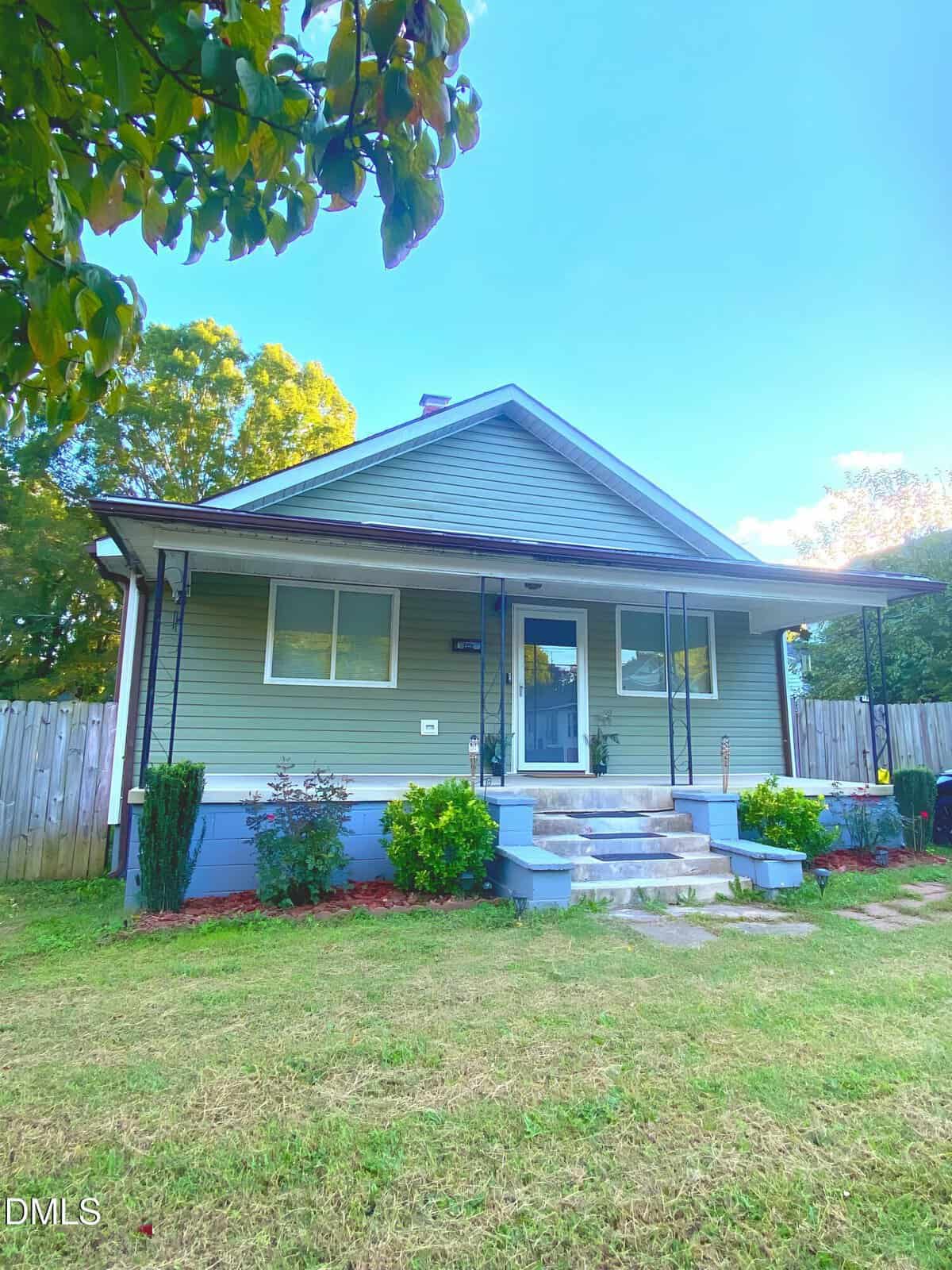 A single-story house at 306 Chavasse Avenue in Henderson features light gray siding, a small covered front porch, blue steps, and white trim. A few shrubs and plants accent the grassy yard, with a wooden fence in the background.