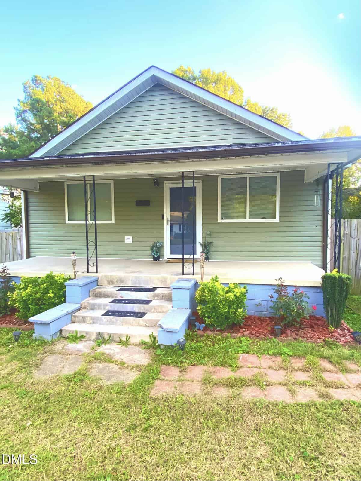 A single-story house with light green siding, a covered front porch supported by metal posts, blue steps, and a small garden with bushes in front. There are two windows and a blue front door.