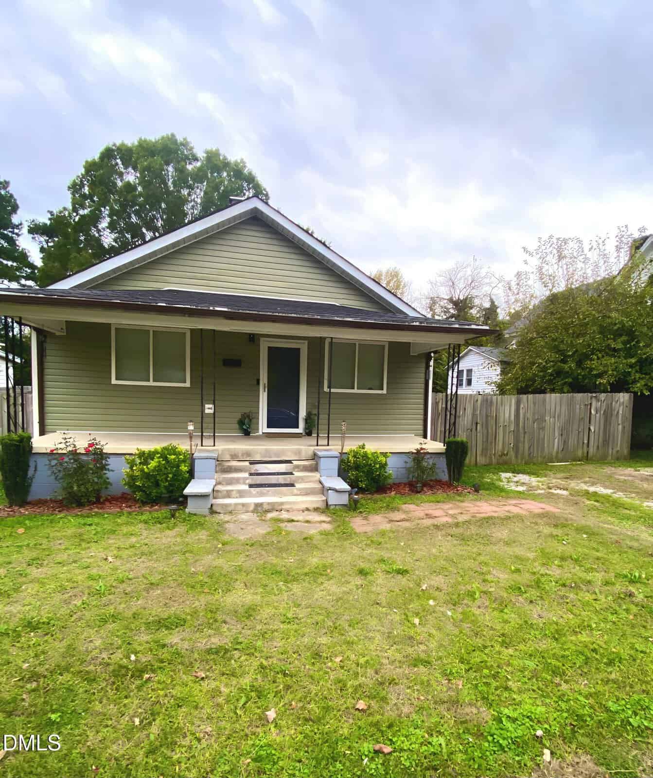 Single-story house with light green siding, white trim, and a covered front porch. There are bushes in front, concrete steps leading to the porch, and a driveway with a wooden fence on the right.