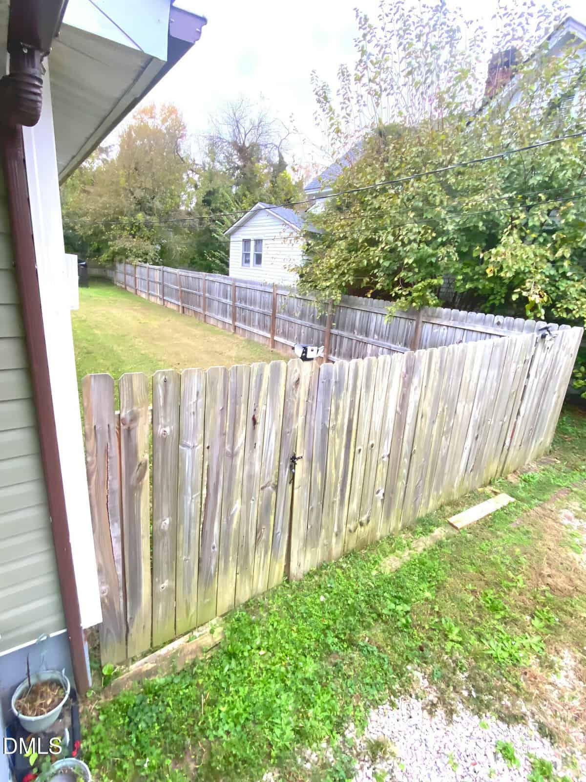 A wooden privacy fence encloses a backyard with grass. The fence is weathered and has a gate. There are trees and another house beyond the fence, and the side of a house is visible in the foreground.