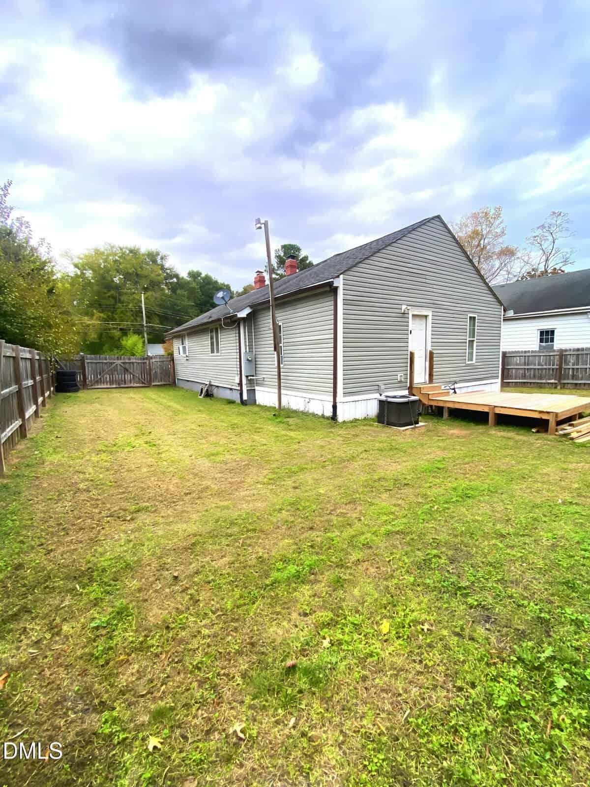 A small gray house with white trim sits in a fenced backyard with a grassy lawn, wooden deck, and central air unit. The sky is overcast and neighboring houses are visible on either side.