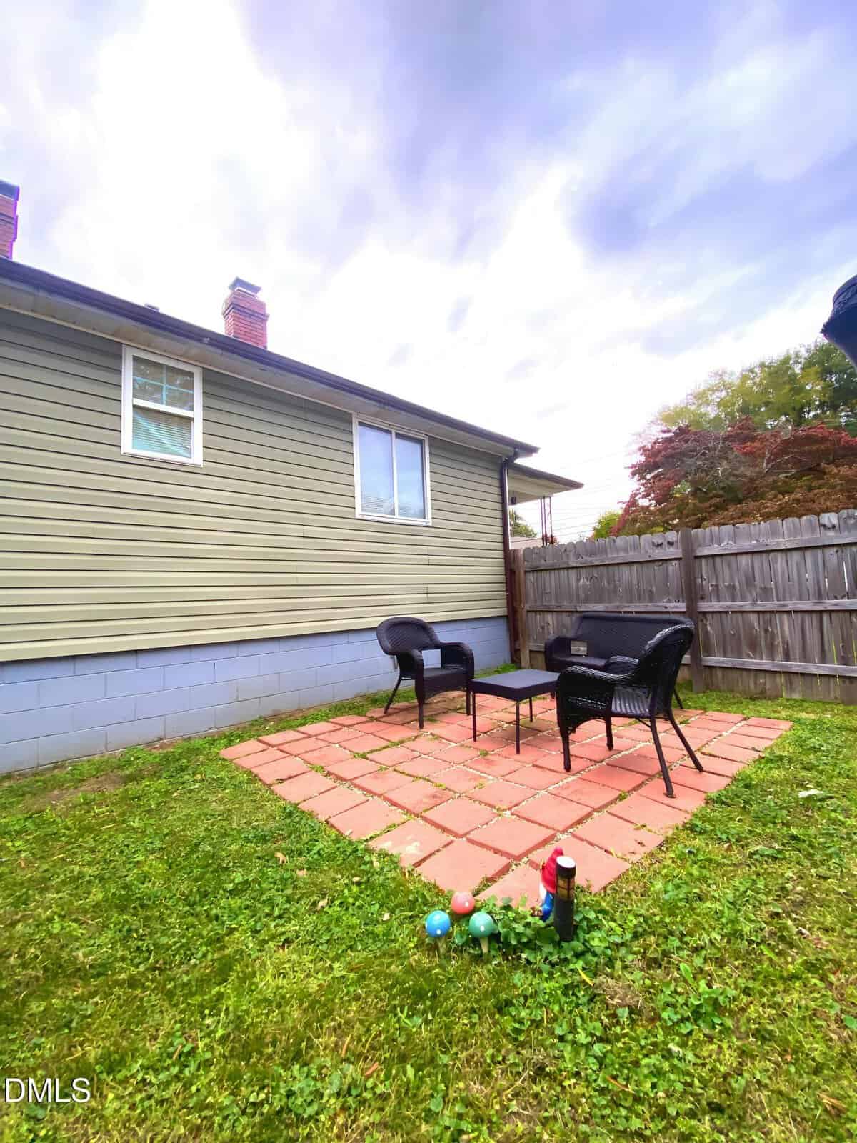 A small backyard patio with red square pavers, two black wicker chairs, a matching table, and a wooden fence. Lawn surrounds the patio, and a few bocce balls and a drink can are on the grass nearby.