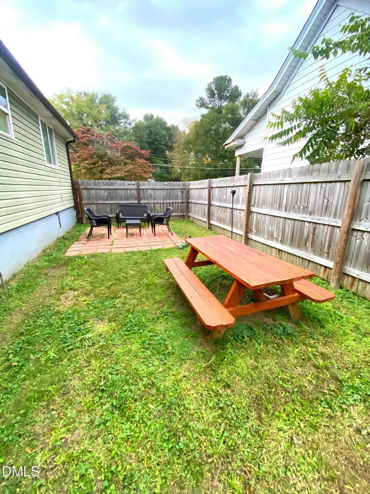 A backyard with a wooden picnic table on grass, a paved patio area with a table and chairs, and a wooden fence separating the yard from neighboring houses.
