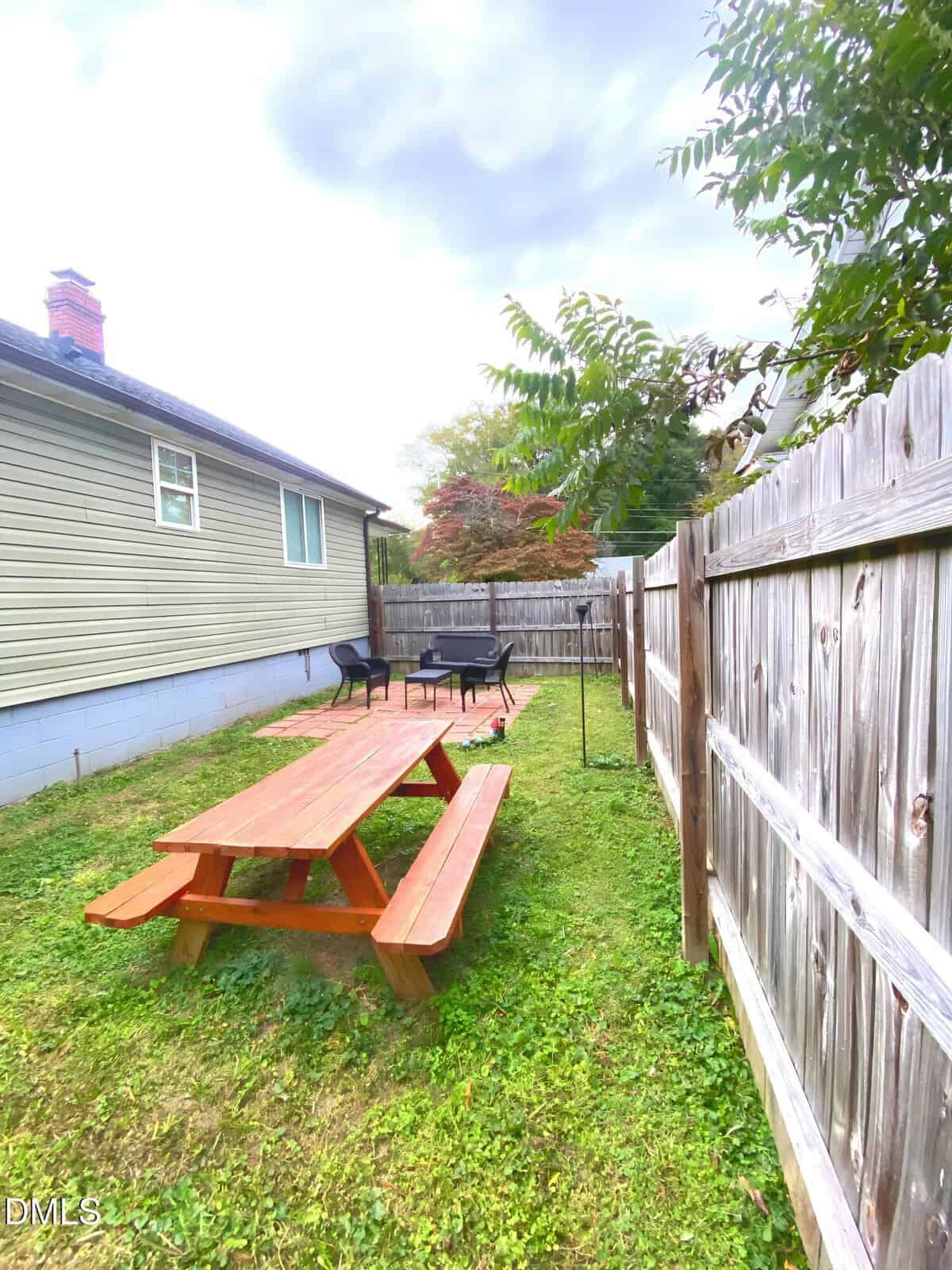 A backyard with a wooden picnic table on grass, two black chairs, a small round table, and a grill near a wooden privacy fence. The side of a house is visible on the left. Trees and cloudy sky are in the background.