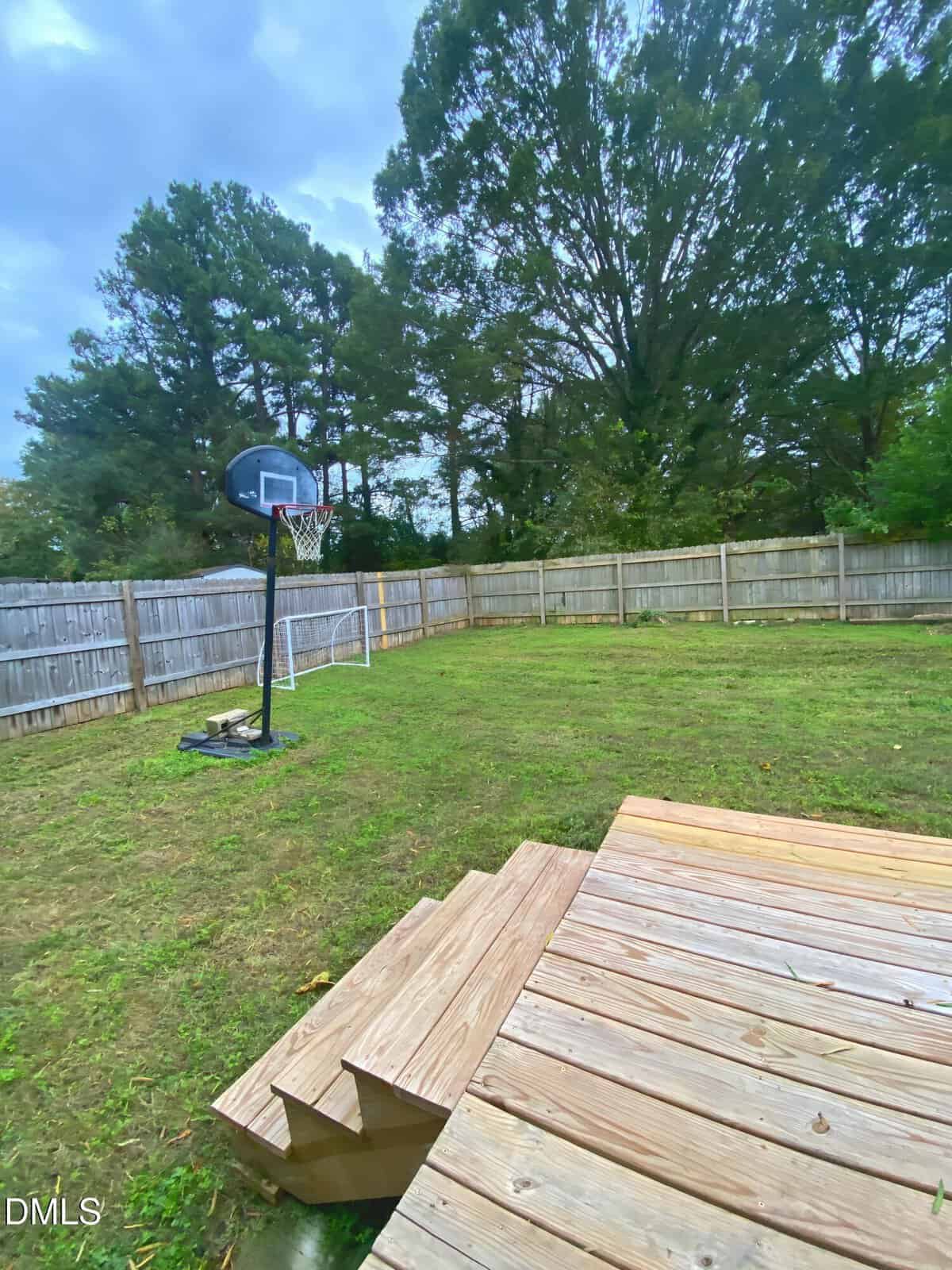 A backyard with a wooden deck, several wooden steps leading to the grass, a basketball hoop on the left, and a wooden privacy fence enclosing the area. Tall trees are visible behind the fence under a cloudy sky.