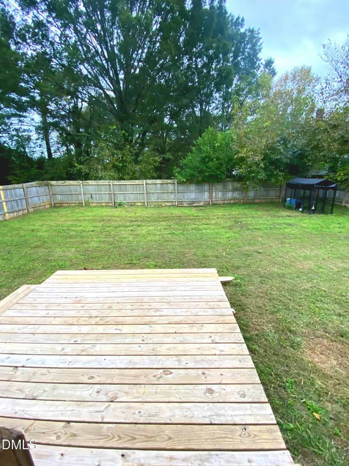 A wooden deck overlooks a fenced backyard with green grass, a few trees, and a small black structure in the corner.