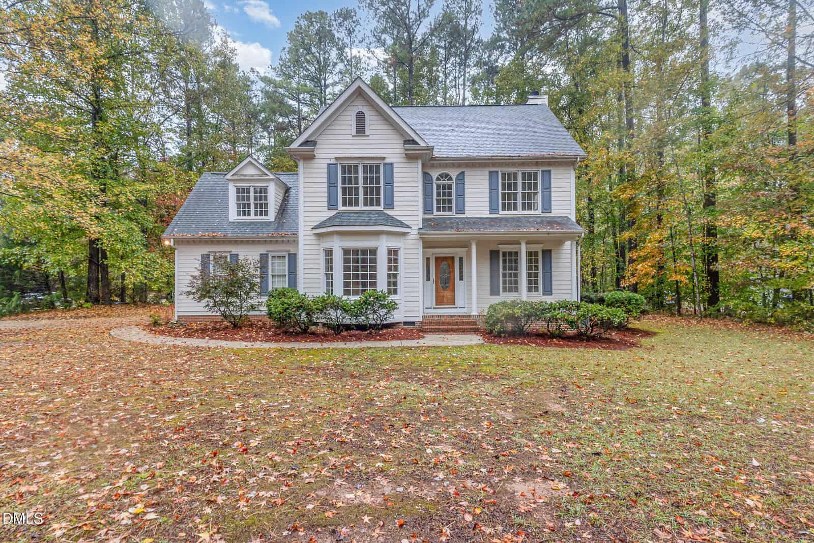 A two-story white house with gray shutters and a covered front porch, surrounded by trees with autumn foliage and a grassy front yard. The sky is partly cloudy.