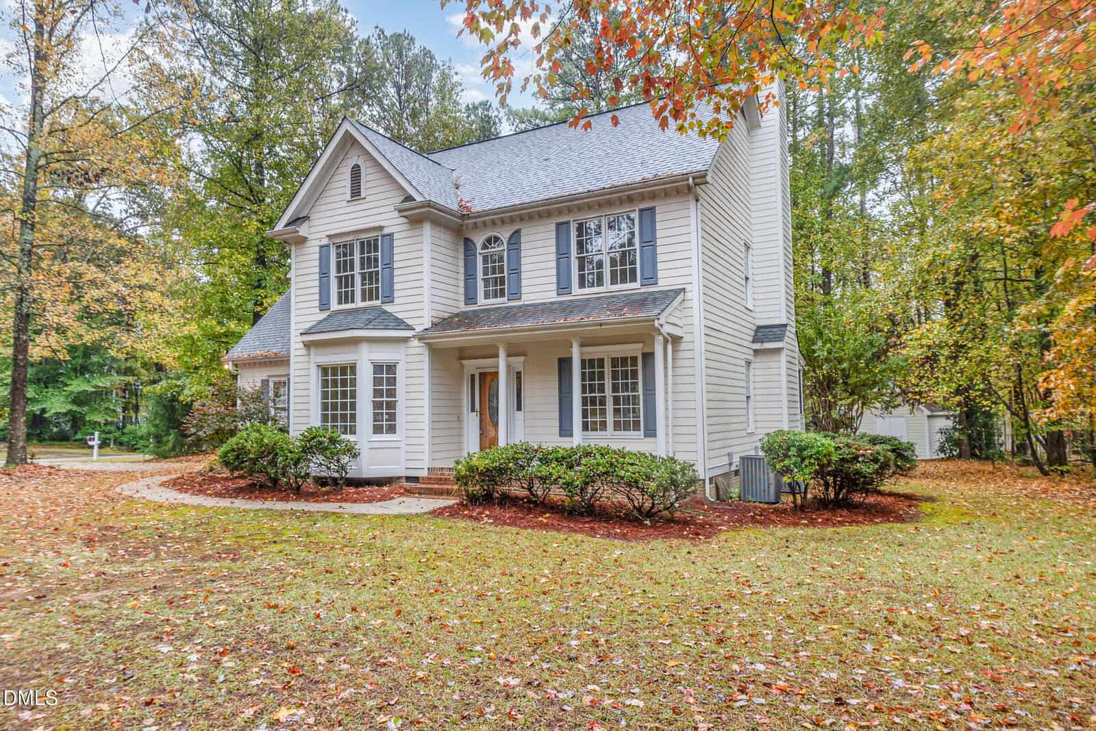 A two-story suburban house with white siding, dark shutters, and a covered front porch, surrounded by trees and autumn foliage. A curved walkway leads to the front door, and shrubs line the front of the house.