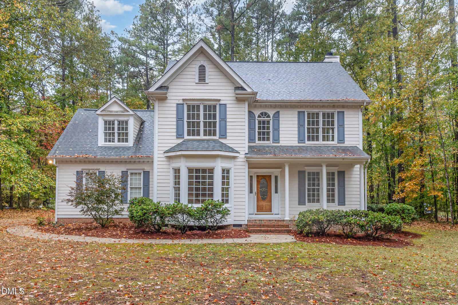A two-story white house with gray shutters and roof at 3333 Keighley Forest Drive, Wake Forest, is surrounded by trees. It features a bay window, a welcoming front porch, wooden door, and a curved path bordered by shrubs and fresh mulch.