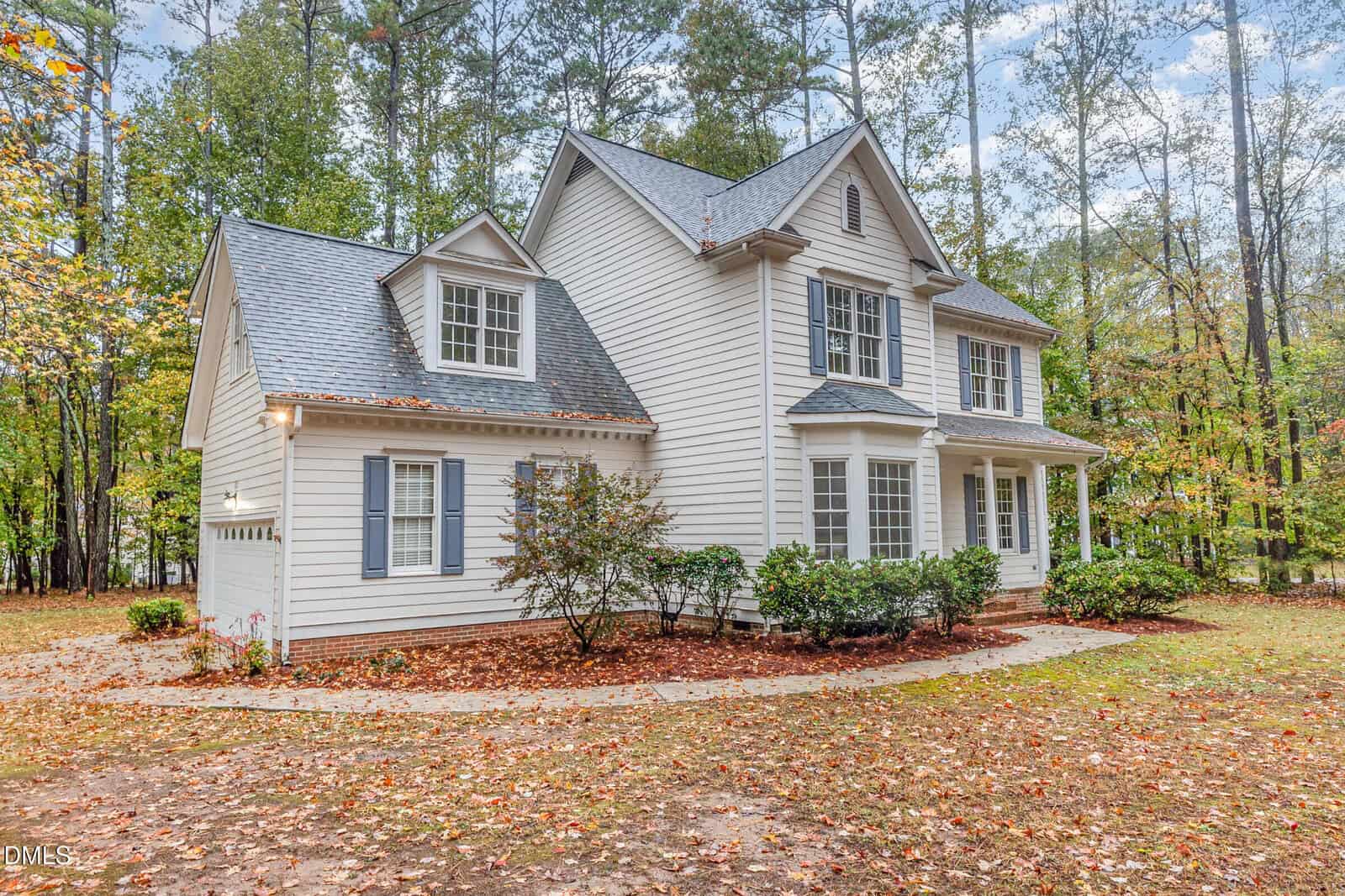 Two-story white house with gray shutters, gabled roof, and a front porch, surrounded by trees and a yard covered in fallen autumn leaves.