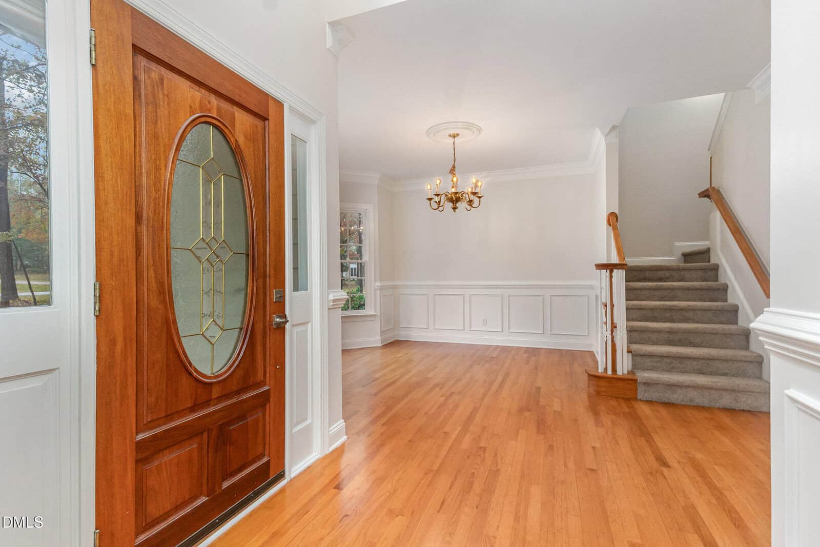 A bright entryway with a wooden front door, hardwood floors, white wainscoting, a chandelier, and carpeted stairs leading up. Large windows let in natural light.