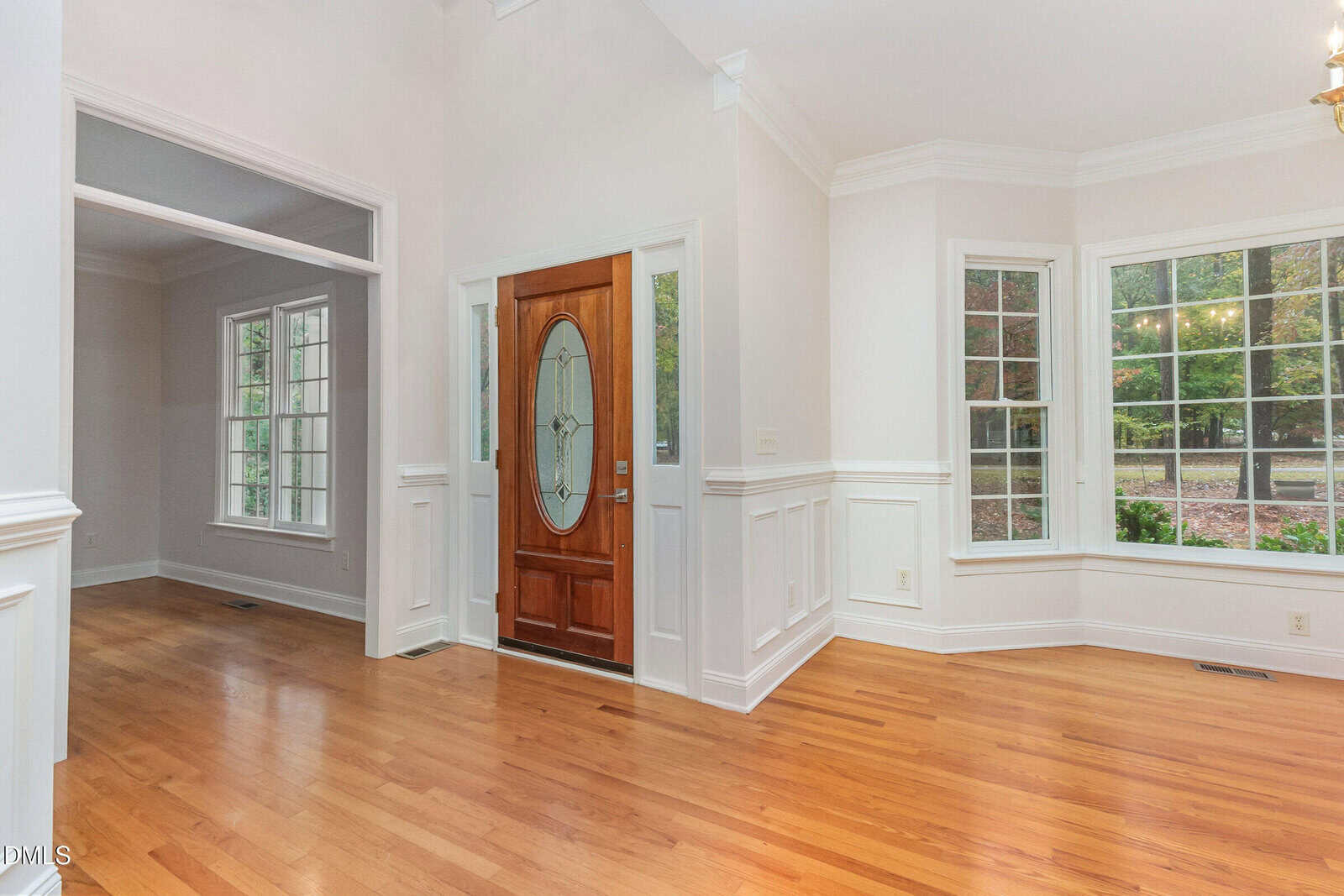 A bright entryway with hardwood floors, white wainscoting, a wooden front door with an oval glass insert, and large windows overlooking greenery outside.