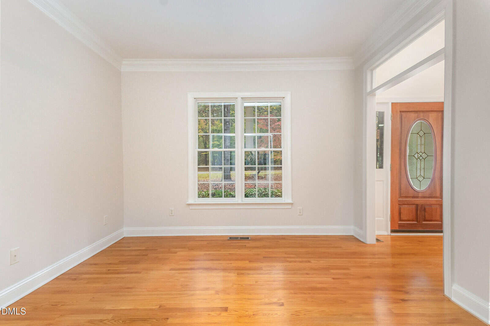 A bright, empty room with light-colored walls, wood flooring, a large window, and an open doorway leading to an entryway with a wooden front door featuring an oval glass design.