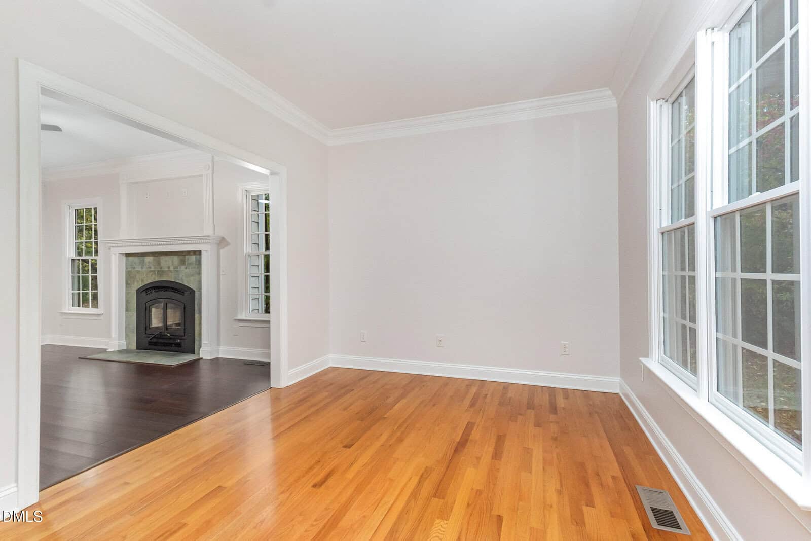 A bright, empty room with light-colored walls and a hardwood floor, large windows on the right, and an open doorway leading to a room with dark flooring and a fireplace.
