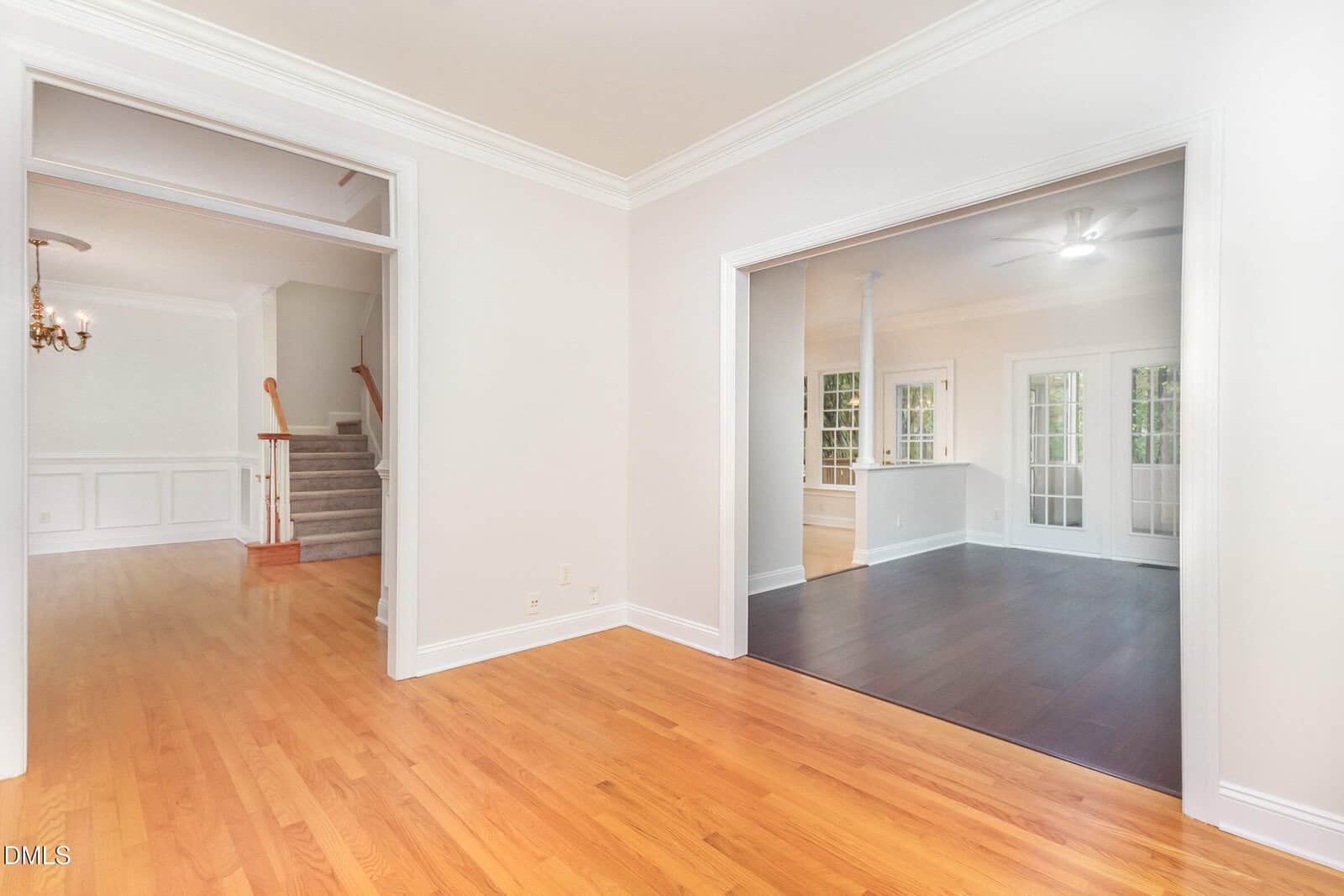 Bright, empty room with light wood flooring, white walls, and wide entryways leading to a staircase with a chandelier and a darker-floored sunroom with large windows and French doors.