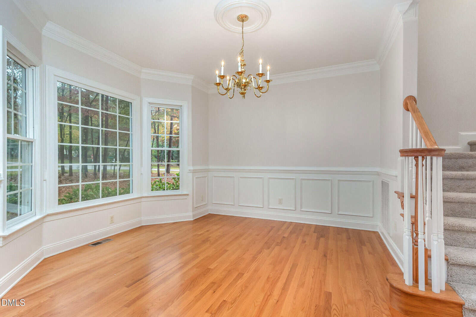 A bright, empty room with large windows, hardwood floors, white wainscoting, a gold chandelier, and a carpeted staircase leading upward on the right. Trees are visible outside through the windows.