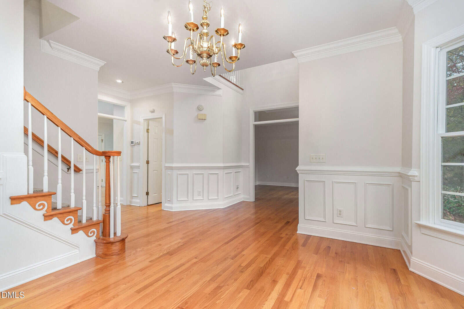 Bright foyer with hardwood floors, white walls, crown molding, a gold chandelier, wooden staircase with white railing, and large window. Wainscoting accents the walls, and doors lead to other rooms.