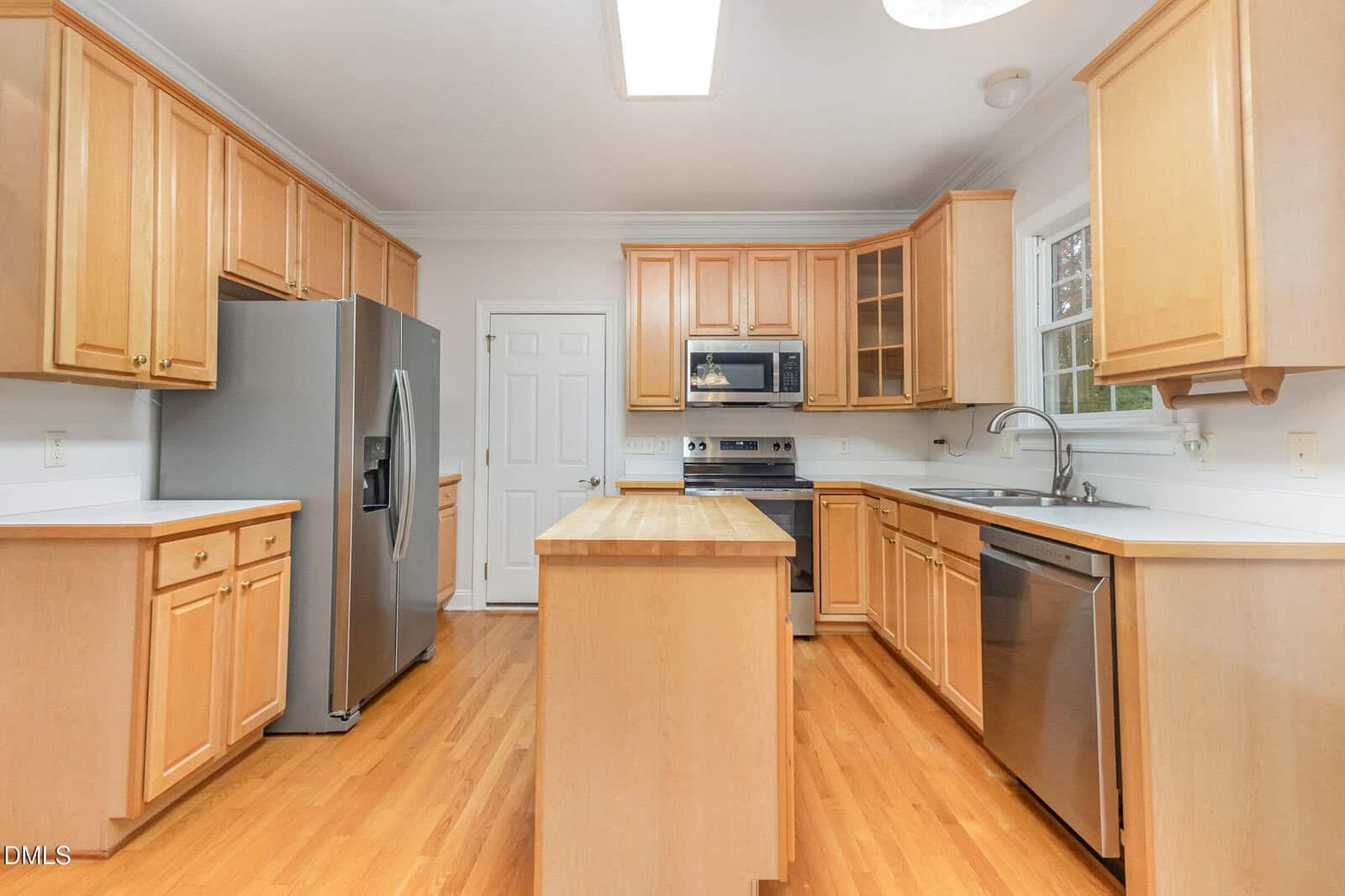Modern kitchen with light wood cabinets, stainless steel appliances, a central island, white countertops, and hardwood floors. A window is above the sink, and a door is in the background.