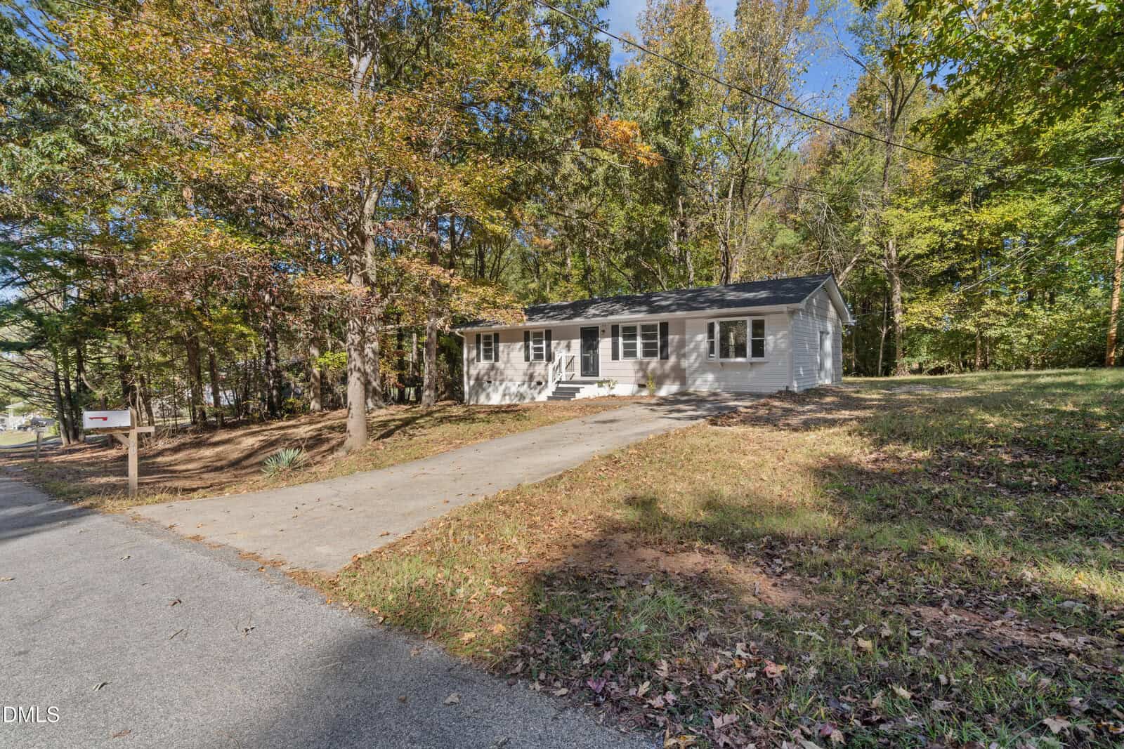 A single-story white house with black shutters and a gray roof, set back from the road with a concrete driveway and surrounded by trees with autumn leaves. A mailbox is visible near the road.