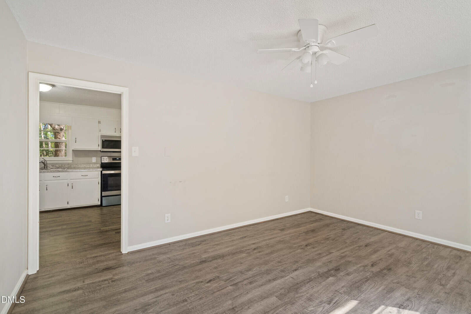 A room with light-colored walls, wood-look flooring, a white ceiling fan, and an open doorway leading to a kitchen with white cabinets and stainless steel appliances.