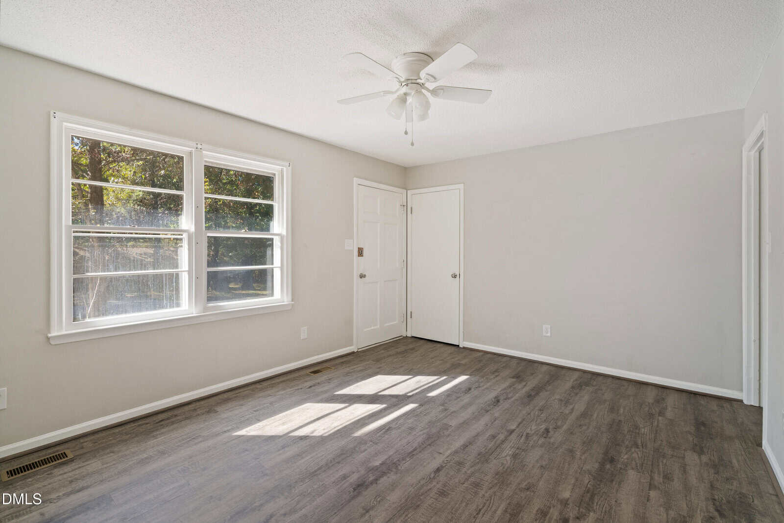 Empty room with light gray walls, a white ceiling fan, two white doors, and a large window letting in sunlight. The floor is dark wood-look laminate. The room is clean and undecorated.