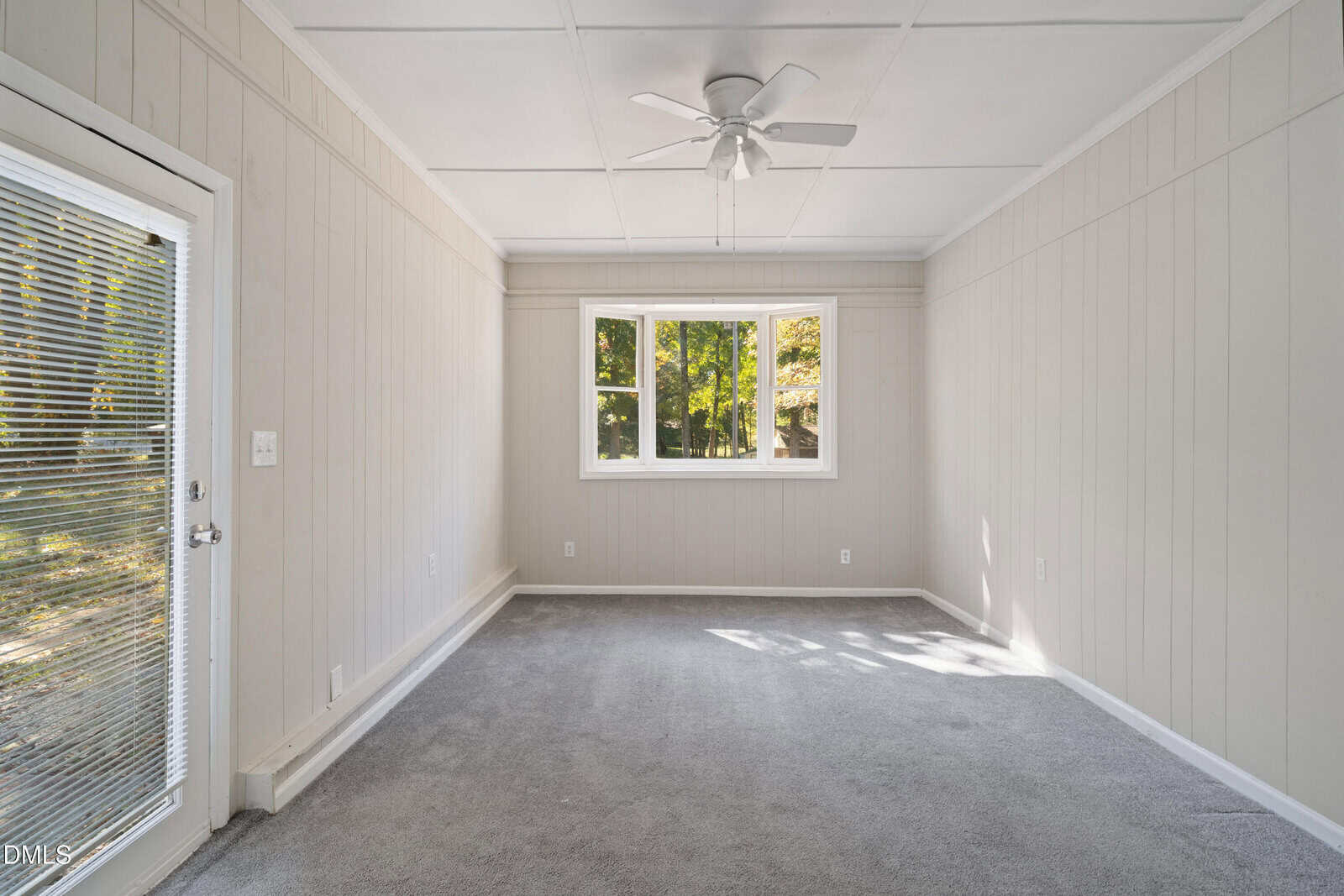 A small, empty room with light grey carpet, beige vertical panel walls, a white ceiling with a ceiling fan, a door with blinds on the left, and three windows showing green trees outside.