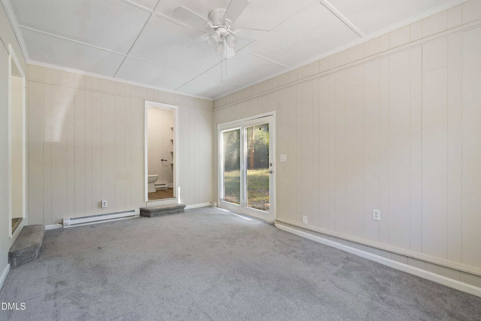 A carpeted room with light-colored wood paneling, a ceiling fan, sliding glass doors to the outside, a small window, and an open door leading to a bathroom.
