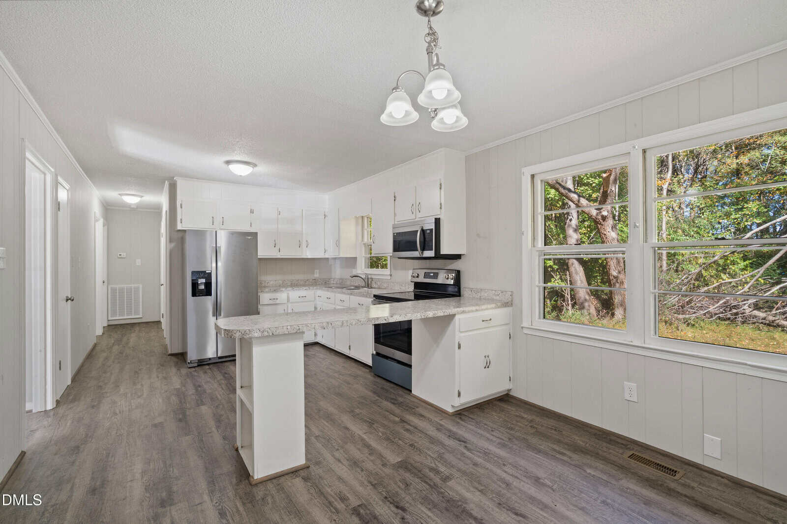 A kitchen with white cabinets, stainless steel appliances, marble-pattern countertops, and wood-look flooring. There is a large window by the dining area, letting in natural light and showing trees outside.