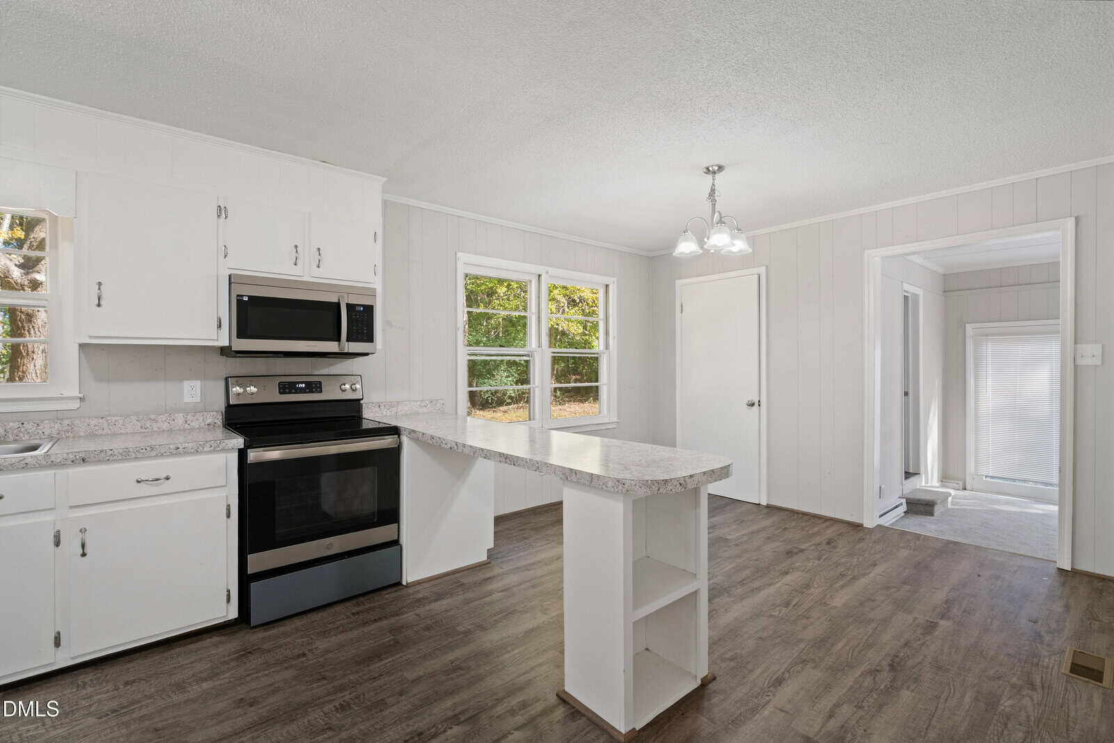 A kitchen with white cabinets, stainless steel appliances, a marble-patterned peninsula, wood-look flooring, and large windows letting in natural light. A door and a light fixture are also visible in the space.
