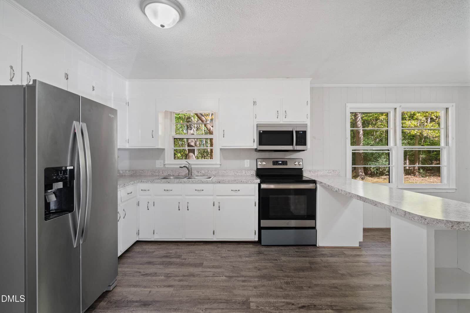 A kitchen with white cabinets, stainless steel appliances including a refrigerator, oven, and microwave, a double sink, marble-patterned countertops, and large windows overlooking greenery. The floor is dark wood.
