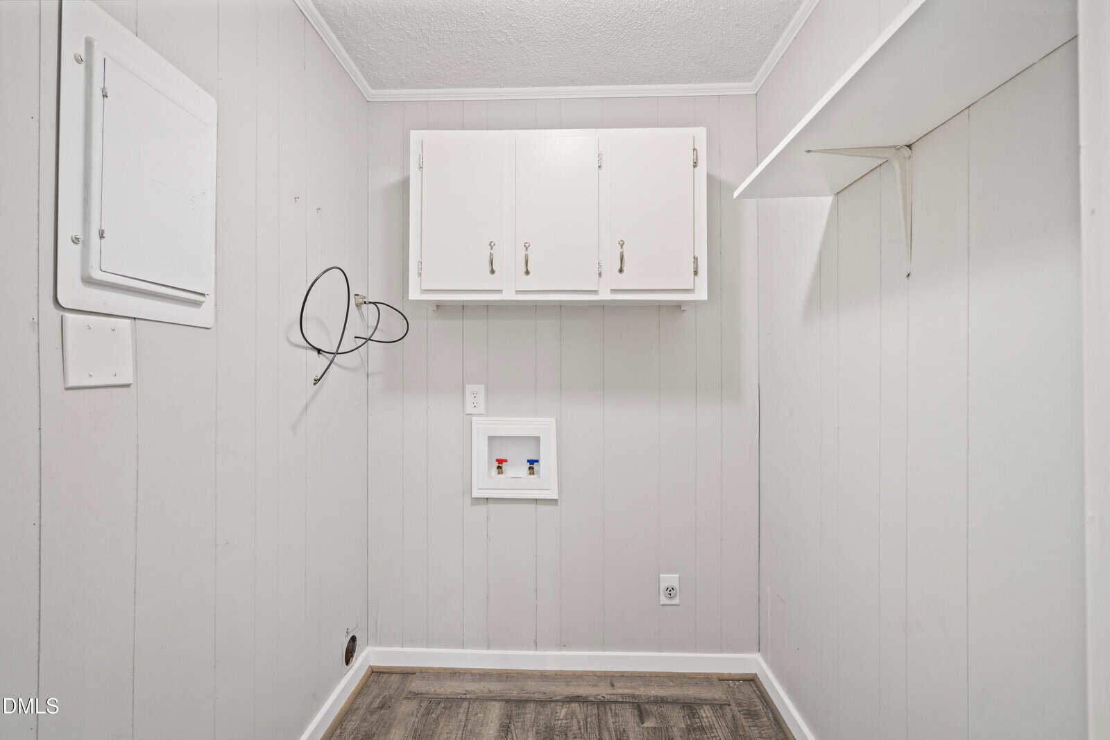 Small laundry room with light gray paneled walls, white cabinets, hookups for washer and dryer, exposed wires, and wood-style flooring. The room appears empty and clean.