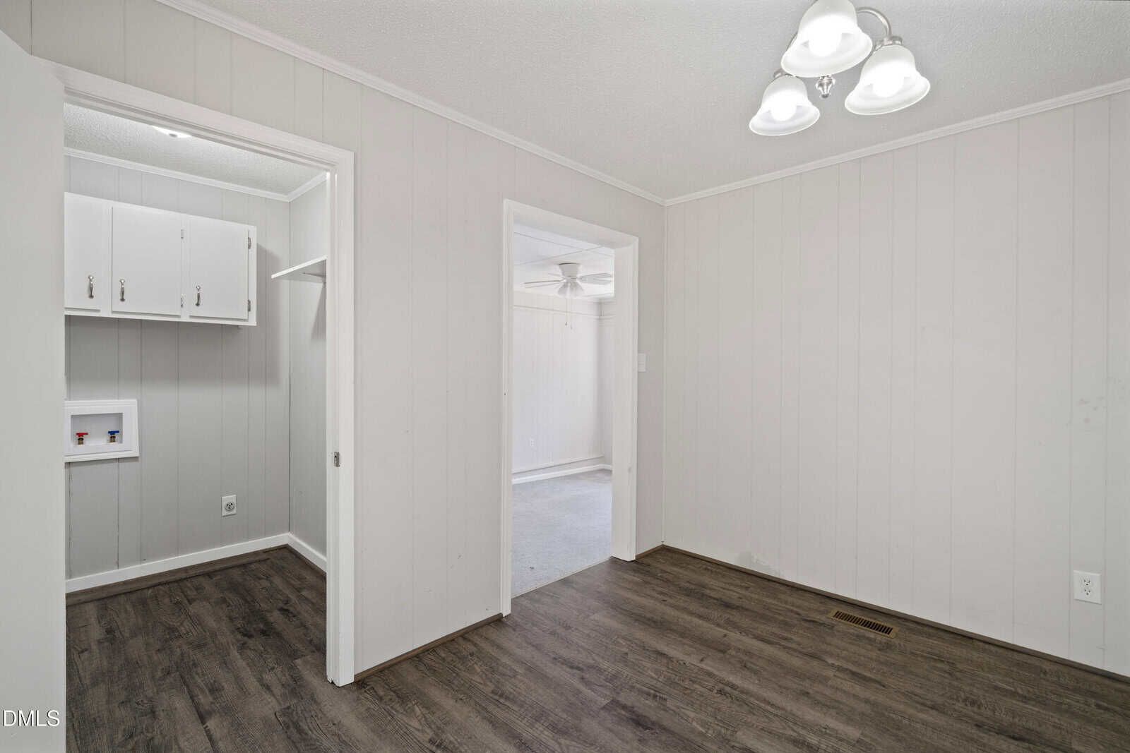 A small room with wood flooring, light gray paneled walls, a ceiling light fixture, and doorways leading to a laundry area and another room with carpet.