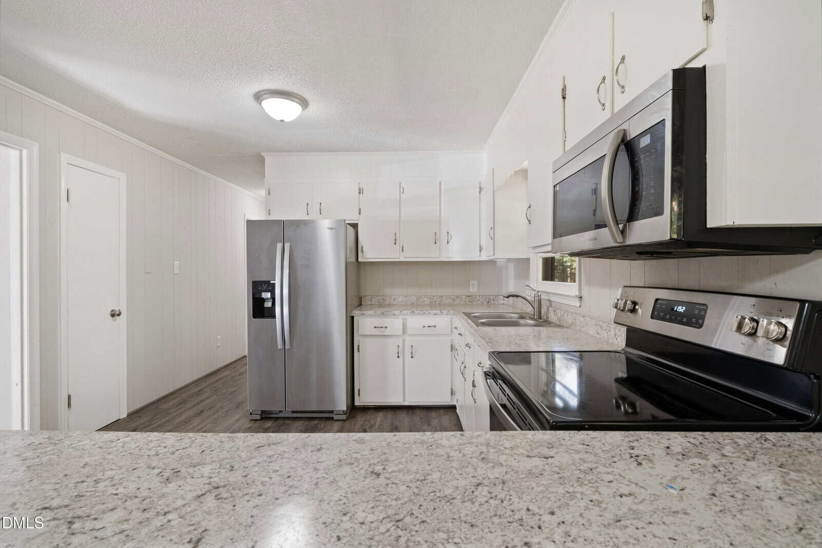 A modern kitchen with white cabinets, stainless steel appliances including a refrigerator, oven, and microwave, light-colored countertops, and wood-look flooring under a ceiling light.