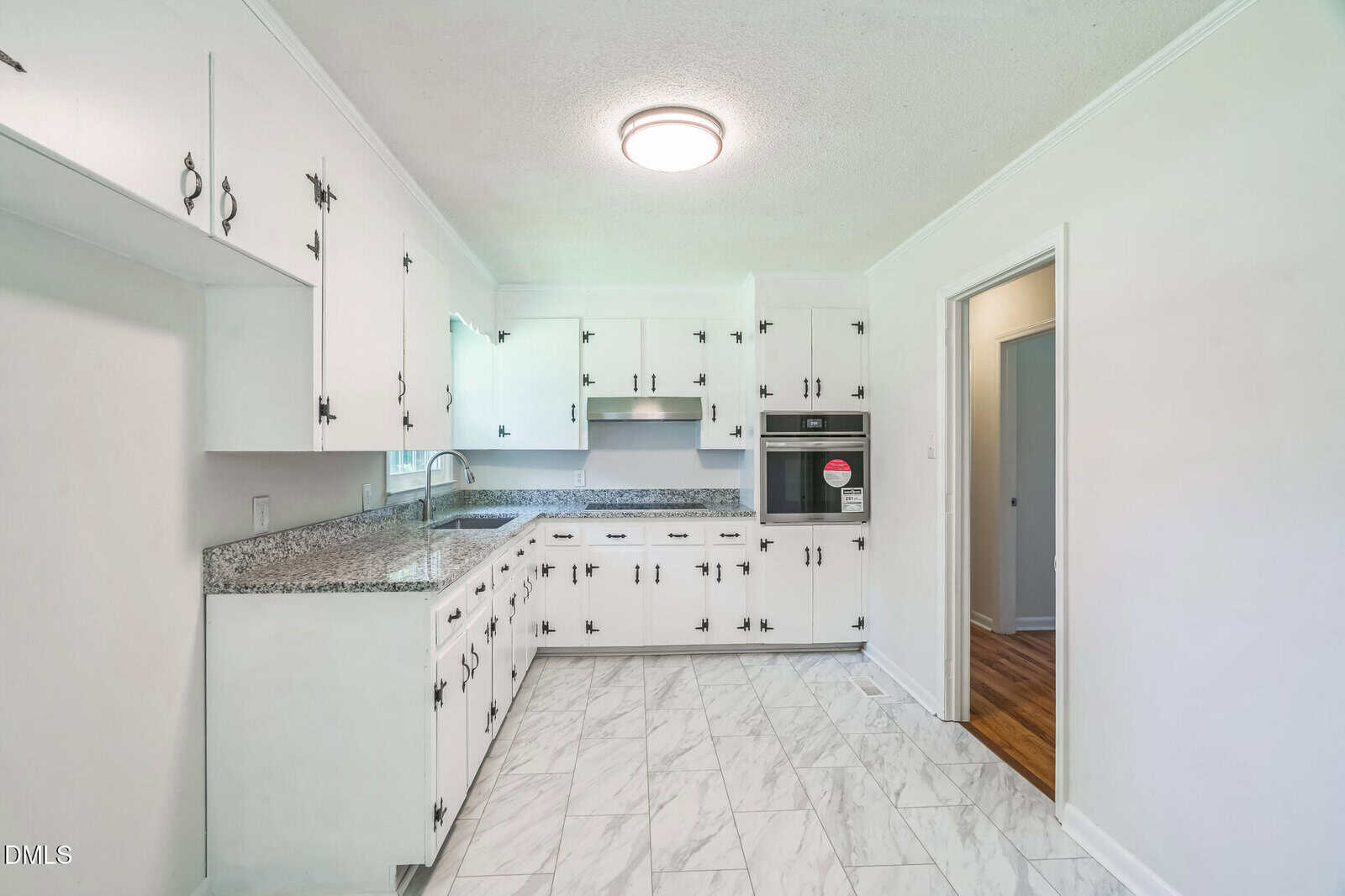 A kitchen with white cabinets, silver handles, granite countertops, and a stainless steel oven. The floor is tiled in white with gray veins, and there is a circular ceiling light. An open doorway leads to another room.