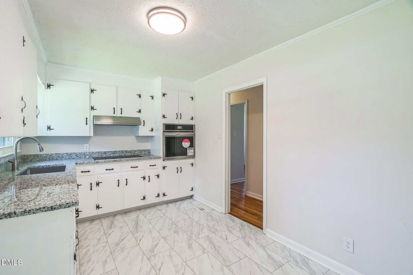 A kitchen with white cabinets, granite countertops, stainless steel appliances, and a tile floor. There is a doorway leading to another room with wooden flooring. The walls are light-colored and there is a ceiling light fixture.