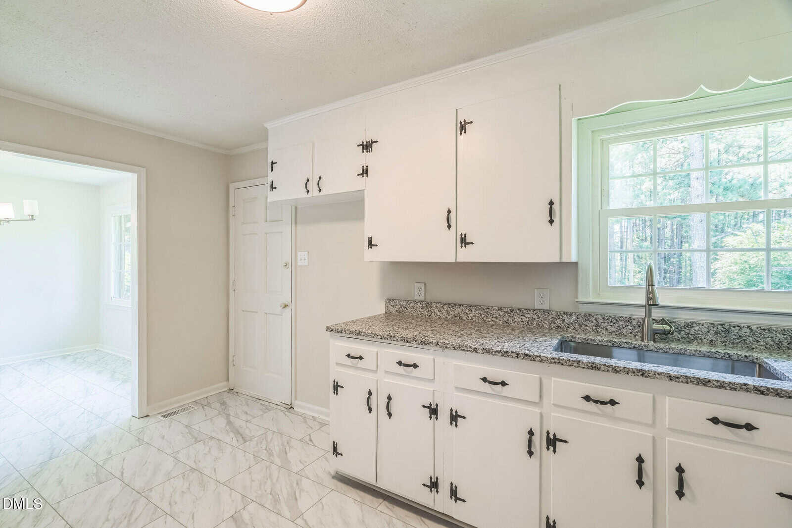 Bright kitchen with white cabinets, black hardware, granite countertop, stainless steel sink, and large window. White tile floor and doorway opening to a sunlit dining area with a chandelier.