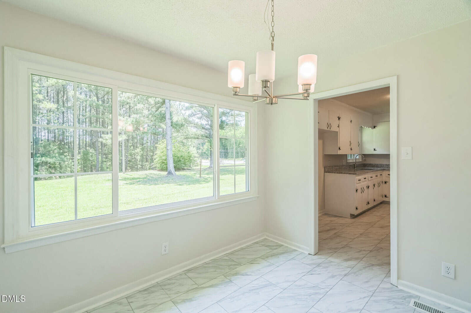 Bright dining area with large windows, white walls, and tiled floor, featuring a modern ceiling light. A doorway leads to a white kitchen with cabinets and countertops. Trees and grass are visible outside the window.